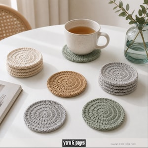 Könnte beinhalten: Eine Sammlung handgefertigter Häkeluntersetzer in verschiedenen neutralen Farben, darunter Beige, Grau und Hellbraun, auf einem weißen Tisch angeordnet. Eine Tasse Tee steht auf einem grünen Untersetzer. Die Untersetzer haben ein Spiraldesign.