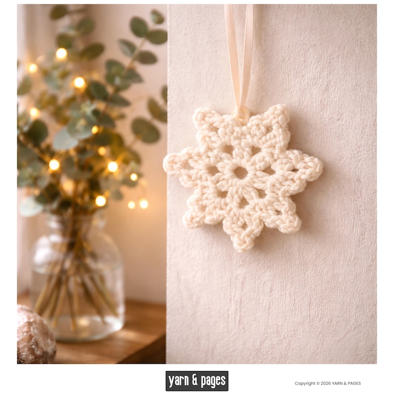 May include: A cream-colored, crocheted snowflake ornament with a delicate, openwork design. The snowflake is suspended by a cream-colored ribbon. The background is a neutral, textured wall. The image also includes a blurred vase of greenery and fairy lights.