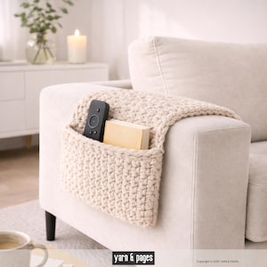 Puede incluir: Un bolsillo de brazo de sofá de ganchillo color crema, con un mando a distancia y un libro pequeño dentro. El bolsillo está colocado sobre el brazo de un sofá beige. El fondo incluye una vela encendida y un jarrón con vegetación.
