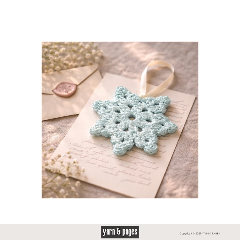 May include: A light blue crocheted snowflake ornament with a cream-colored ribbon, resting on a vintage-style card. The snowflake has a delicate, openwork design. A sealed envelope and sprigs of white flowers are in the background.
