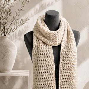 May include: A cream-colored, chunky knit scarf with a waffle-like texture. The scarf is draped around a dark gray mannequin torso, showcasing the open, grid-like stitch pattern and the soft, thick yarn construction of the accessory.
