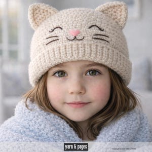 Puede incluir: Un gorro de gato de ganchillo color crema con rasgos faciales bordados, que incluyen una nariz rosa y bigotes negros. El gorro tiene dos orejas puntiagudas y un borde acanalado. Una persona joven lleva el gorro, combinado con una prenda azul claro.