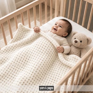 Könnte beinhalten: Ein Baby liegt in einem Holzbettchen und ist mit einer cremefarbenen, strukturierten Decke bedeckt. Ein Teddybär sitzt neben dem Baby. Die Decke hat ein gewebtes Muster. Der Text "yarn & pages" befindet sich unten im Bild.