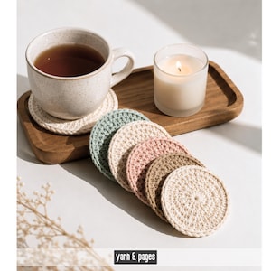 May include: A collection of handmade crochet coasters in various pastel colors, including cream, blue, and pink. A mug of tea sits on a coaster, next to a lit candle and a wooden tray. The text "yarn & pages" is at the bottom.