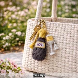 Könnte beinhalten: Ein handgefertigter Häkel-Schlüsselanhänger mit einer Champagnerflasche und einem Glas. Die Flasche ist dunkelbraun mit gelbem Deckel und dem Wort "JOY". Das Glas ist grau und gelb. Der Schlüsselanhänger ist an einer gewebten Tasche mit einem Schlüssel befestigt.