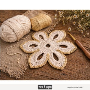 Può includere: Un sottobicchiere a forma di fiore all'uncinetto, color crema, con un bordo dorato, è appoggiato su una superficie di legno. Sono visibili anche due gomitoli di lana e un uncinetto. L'immagine include il testo "yarn & pages".