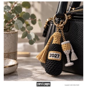 Può includere: Un portachiavi all'uncinetto composto da una bottiglia nera con la parte superiore beige e un'etichetta con la scritta 2027, accompagnato da una nappa beige abbinata e un piccolo ciondolo a forma di flûte da champagne con stelo bianco.