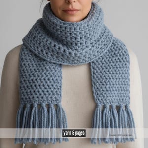 Könnte beinhalten: Hellblauer Häkelschal mit dickem, strukturiertem Gewebe. Der Schal hat einen breiten Kragen und lange, gefranste Enden. Das Bild enthält den Text "yarn & pages".