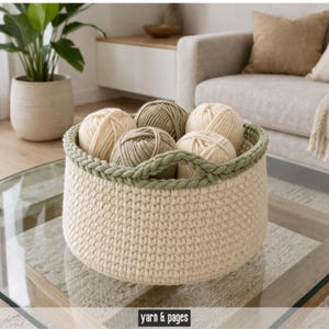 Puede incluir: Una cesta de ganchillo de color crema con un borde trenzado verde, llena de ovillos de lana en tonos crema y verde. El texto "yarn & pages" est&aacute; en la parte inferior de la imagen. La cesta est&aacute; sobre una mesa de cristal.