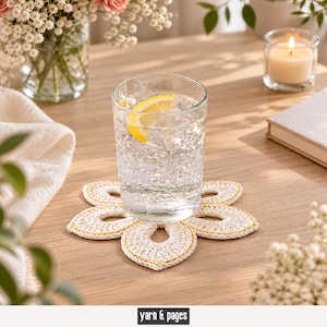 Könnte beinhalten: Ein Glas Wasser mit Zitrone steht auf einem weißen und goldenen Häkelblumen-Untersetzer. Der Untersetzer befindet sich auf einer Holzoberfläche, im Hintergrund eine brennende Kerze und Blumen. Der Text "yarn & pages" steht unten.
