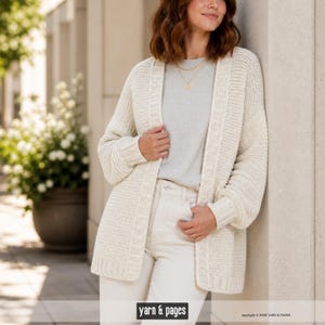 Peut inclure: Cardigan crème à devant ouvert avec un motif en tricot texturé. Le pull à manches longues a une coupe décontractée et est porté sur un haut gris et un pantalon blanc. L'image comprend le texte "yarn & pages".