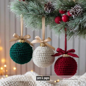 Könnte beinhalten: Drei gehäkelte Weihnachtskugeln in Grün, Weiß und Rot, jede mit einer goldfarbenen Kappe und einem Satinband zum Aufhängen. Die Ornamente hängen an einem festlichen Tannenzweig mit Tannenzapfen und roten Beeren. Der Text "yarn & pages" ist sichtbar.
