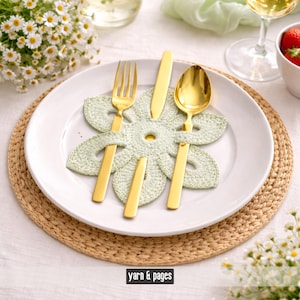 Könnte beinhalten: Ein Gedeck mit einem weißen Teller, goldenem Besteck und einem grünen, gehäkelten blütenförmigen Platzset. Das Bild zeigt einen Blumenstrauß und den Text "yarn & pages".