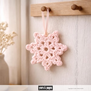 May include: A light pink crocheted snowflake ornament hanging from a wooden peg. The snowflake is made of yarn and has a delicate, openwork design. A light pink ribbon is used for hanging. The background is a light-colored wall.