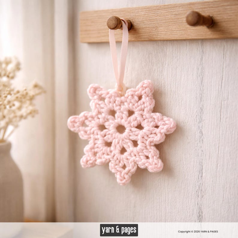 May include: A light pink crocheted snowflake ornament hanging from a wooden peg. The snowflake is made of yarn and has a delicate, openwork design. A light pink ribbon is used for hanging. The background is a light-colored wall.