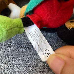 K&ouml;nnte beinhalten: Ein Pl&uuml;schtier aus rotem, gr&uuml;nem und schwarzem Stoff mit einem wei&szlig;en Etikett, auf dem steht: MADE EXCLUSIVELY FOR The Disney Company &copy; DISNEY The Disney Store, Inc. MA-1336(KR)/PA-5501(KR)/CHIO PA 1378: CE.