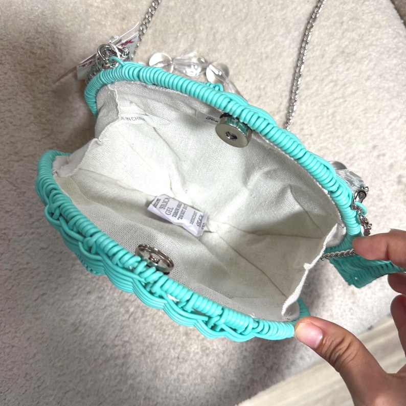 Puede incluir: Un bolso turquesa en forma de concha con una correa de cadena plateada. El bolso tiene una textura tejida alrededor de los bordes y un interior de tela blanca. Un cierre plateado asegura la abertura del bolso. Cuentas transparentes adornan la cadena.