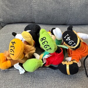K&ouml;nnte beinhalten: Drei Pl&uuml;schfiguren mit eingestickten Jahreszahlen auf den F&uuml;&szlig;en. Eine hat einen gelben Fu&szlig; mit 1934, eine einen gr&uuml;nen Fu&szlig; mit 1938 und eine einen schwarzen Fu&szlig; mit 1941. Die Figuren tragen bunte Outfits mit Stoffelementen in Rot, Orange und Braun.