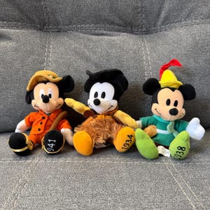 K&ouml;nnte beinhalten: Drei Mickey-Mouse-Pl&uuml;schfiguren in verschiedenen Outfits. Die linke Figur tr&auml;gt einen orangefarbenen Mantel und Hut mit 941 auf dem Fu&szlig;. Die mittlere Figur tr&auml;gt eine braune fellartige Weste mit 1934 auf dem Fu&szlig;. Die rechte Figur tr&auml;gt eine gr&uuml;ne Tunika und einen gelben Hut mit einer roten Feder und 1938 auf dem Fu&szlig;.