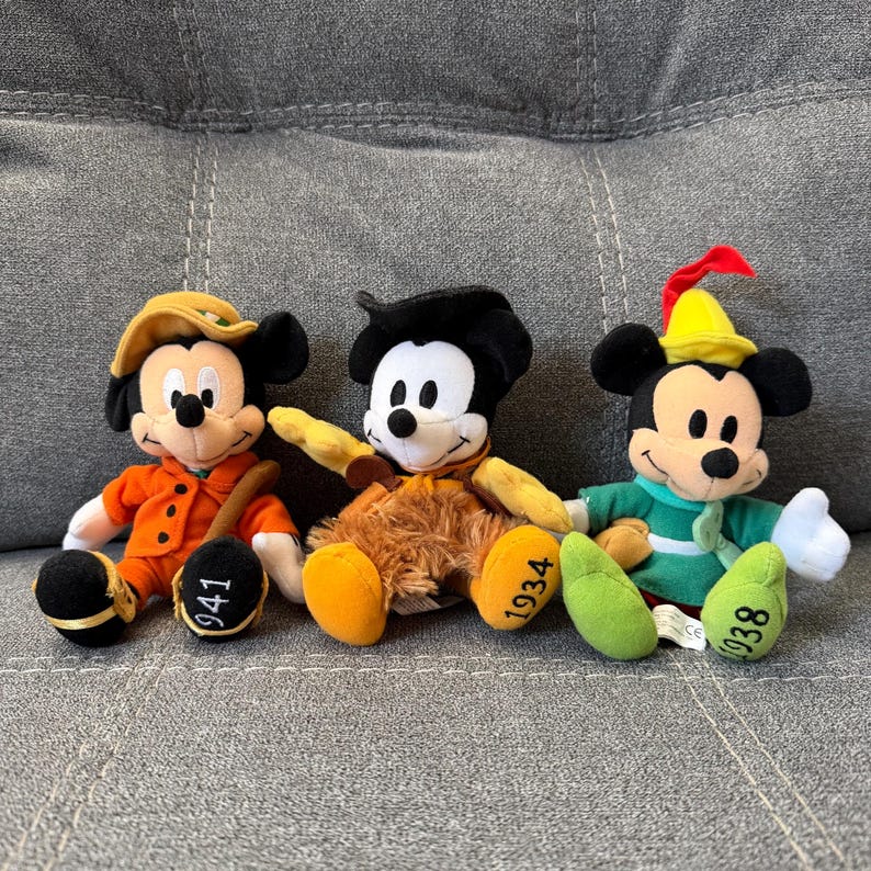 K&ouml;nnte beinhalten: Drei Mickey-Mouse-Pl&uuml;schfiguren in verschiedenen Outfits. Die linke Figur tr&auml;gt einen orangefarbenen Mantel und Hut mit 941 auf dem Fu&szlig;. Die mittlere Figur tr&auml;gt eine braune fellartige Weste mit 1934 auf dem Fu&szlig;. Die rechte Figur tr&auml;gt eine gr&uuml;ne Tunika und einen gelben Hut mit einer roten Feder und 1938 auf dem Fu&szlig;.