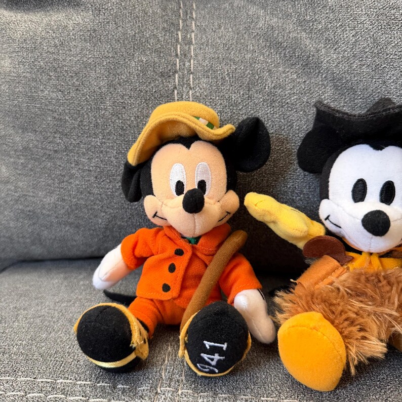 K&ouml;nnte beinhalten: Zwei Pl&uuml;schtiere, die Mickey-Mouse-Figuren darstellen. Eines tr&auml;gt eine orangefarbene Jacke, einen gelben Hut und h&auml;lt einen braunen Stab, mit der Zahl 941 auf dem Fu&szlig;. Das andere ist teilweise sichtbar und tr&auml;gt einen schwarzen Hut und ein braunes Outfit.