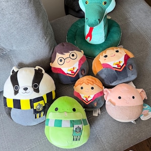 May include: A collection of seven plush toys including a large green snake, a badger wearing a yellow and black scarf, a round character with glasses, a round character with orange hair, a small round character, a green snake with a scarf, and a pink creature.