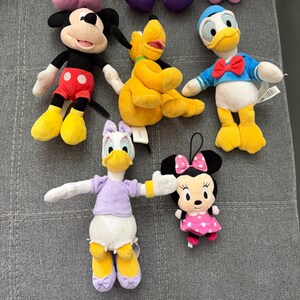 May include: A collection of five plush toys featuring iconic cartoon characters. The set includes a black and red mouse, a yellow dog, a white duck in a blue shirt, a white duck in a lavender top, and a small black mouse with a pink polka dot dress.