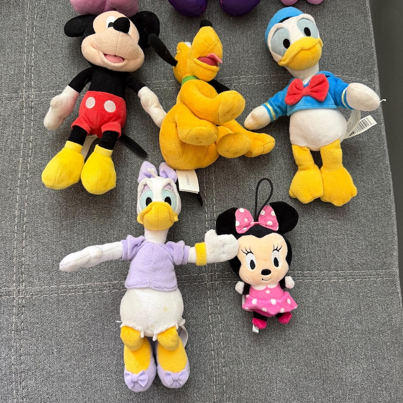 May include: A collection of five plush toys featuring iconic cartoon characters. The set includes a black and red mouse, a yellow dog, a white duck in a blue shirt, a white duck in a lavender top, and a small black mouse with a pink polka dot dress.