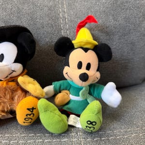 K&ouml;nnte beinhalten: Ein Mickey-Mouse-Pl&uuml;schtier in einer gr&uuml;nen Tunika mit einem gelben Hut, der eine rote Feder tr&auml;gt. Die Figur hat wei&szlig;e Handschuhe, eine braune Tasche an der Taille und hellgr&uuml;ne Schuhe mit der Zahl 938 auf der Sohle des linken Fu&szlig;es.