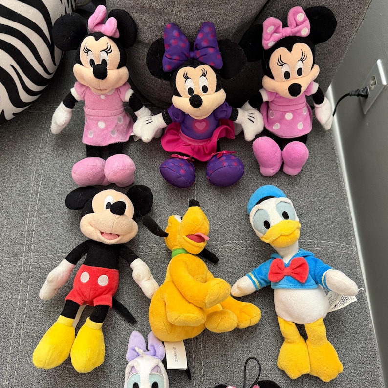 May include: A collection of six plush toys featuring Disney characters including three Minnie Mouse dolls in pink and purple outfits, one Mickey Mouse doll in red shorts, one yellow Pluto dog, and one Donald Duck wearing a blue sailor shirt.