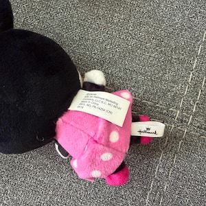May include: A plush toy featuring a black head and a pink dress with white polka dots. A white fabric tag is attached to the back, displaying text including Disney, Hallmark Marketing Company, LLC, and Made in China. A small white ribbon tag with the Hallmark logo is also visible.