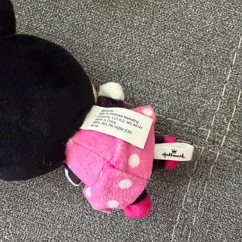 May include: A plush toy featuring a black head and a pink dress with white polka dots. A white fabric tag is attached to the back, displaying text including Disney, Hallmark Marketing Company, LLC, and Made in China. A small white ribbon tag with the Hallmark logo is also visible.