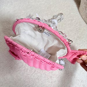 May include: A pink woven fish-shaped handbag with a clear beaded handle. The bag has a white interior and a magnetic clasp. The fish design includes a tail and fins. The bag is open, revealing the interior.