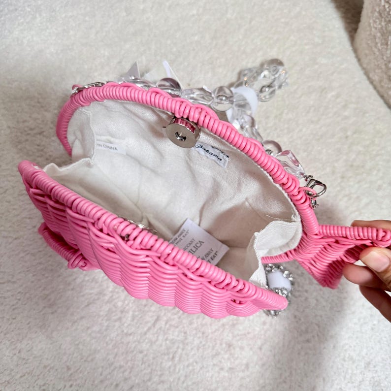 May include: A pink woven fish-shaped handbag with a clear beaded handle. The bag has a white interior and a magnetic clasp. The fish design includes a tail and fins. The bag is open, revealing the interior.