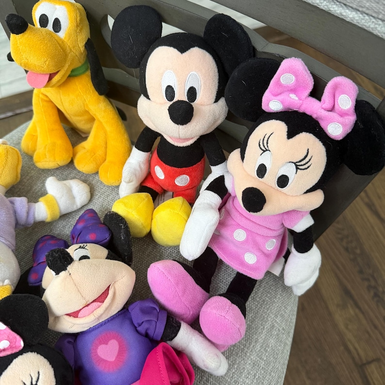 May include: A collection of plush toys featuring Pluto with yellow fur, Mickey Mouse in red shorts, and two Minnie Mouse dolls, one wearing a pink polka-dot dress with a matching bow and the other in a purple dress with a pink heart graphic.