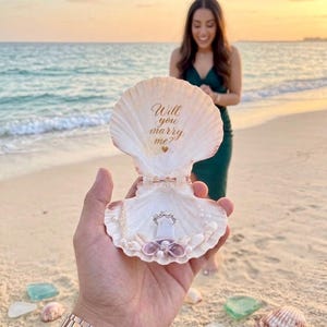 May include: An open white seashell ring holder featuring the gold text Will you marry me? with a small gold heart. The shell is decorated with tiny white pearls and smaller shells, holding a silver ring with multiple stones in the center.