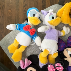 May include: A plush Donald Duck toy wearing a blue shirt with a red bow tie and yellow feet, alongside a plush Daisy Duck toy in a lavender shirt with a matching bow, plus parts of other plush toys including yellow and black patterned fabric.