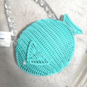 Puede incluir: Un bolso turquesa con forma de pez con un dise&ntilde;o tejido. El bolso tiene una correa de cadena de cuentas transparente y un peque&ntilde;o acento de perla. El bolso est&aacute; hecho de un material tejido y tiene un dise&ntilde;o de cola de pez.