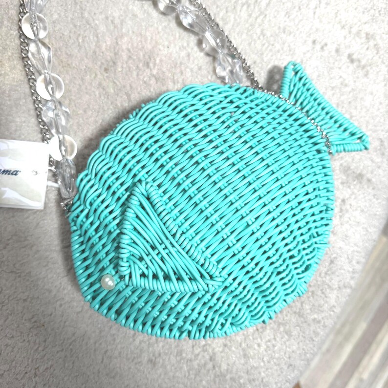 Puede incluir: Un bolso turquesa con forma de pez con un dise&ntilde;o tejido. El bolso tiene una correa de cadena de cuentas transparente y un peque&ntilde;o acento de perla. El bolso est&aacute; hecho de un material tejido y tiene un dise&ntilde;o de cola de pez.