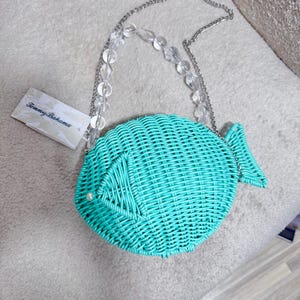 Puede incluir: Un bolso turquesa con forma de pez y textura tejida. El bolso tiene una correa de cadena plateada con detalles de cuentas transparentes. Se adjunta una etiqueta con el logotipo de Tommy Bahama. Una peque&ntilde;a perla est&aacute; en el costado del pez.