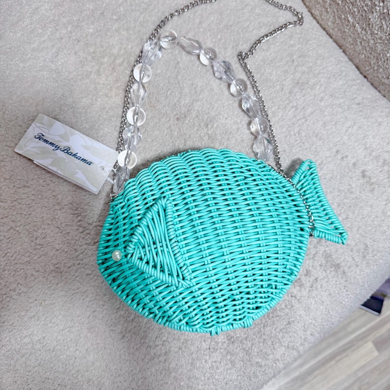 Puede incluir: Un bolso turquesa con forma de pez y textura tejida. El bolso tiene una correa de cadena plateada con detalles de cuentas transparentes. Se adjunta una etiqueta con el logotipo de Tommy Bahama. Una peque&ntilde;a perla est&aacute; en el costado del pez.
