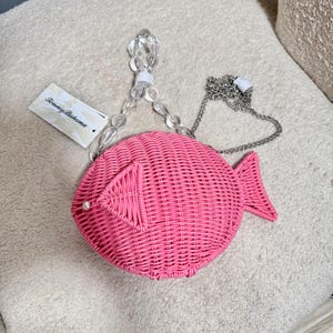 May include: A pink woven fish-shaped handbag with a clear beaded handle and a silver chain strap. The bag has a pearl accent and a tag that reads "Tommy Bahama". The bag is made of woven material.
