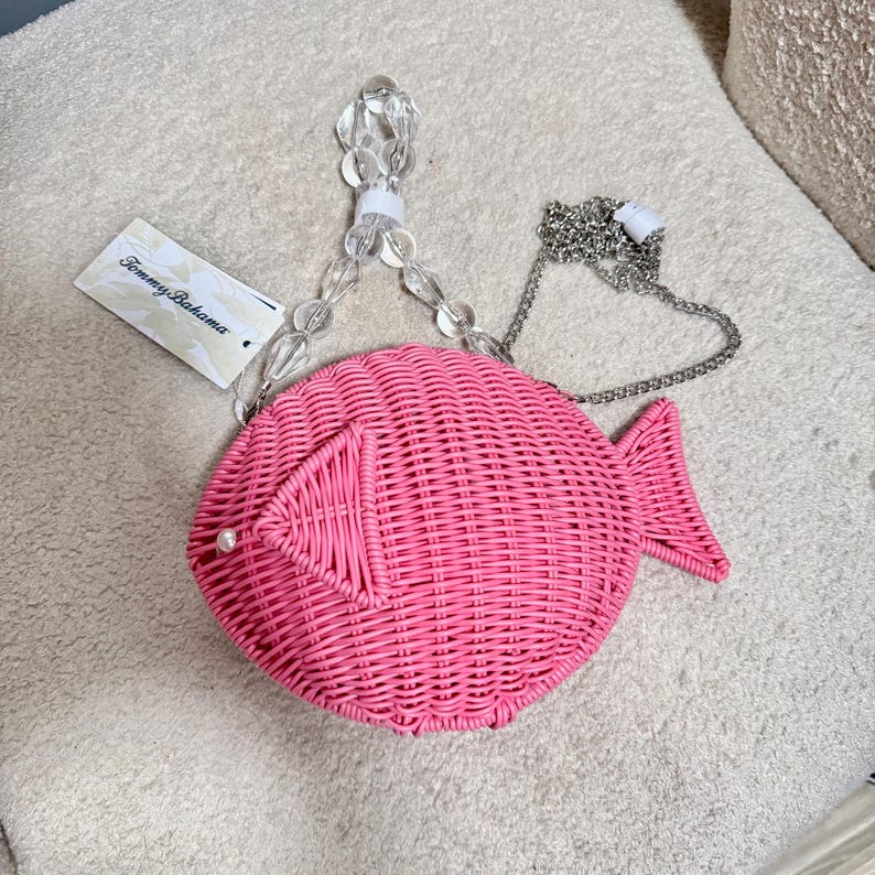 May include: A pink woven fish-shaped handbag with a clear beaded handle and a silver chain strap. The bag has a pearl accent and a tag that reads "Tommy Bahama". The bag is made of woven material.