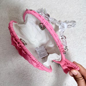 May include: A pink heart-shaped handbag with a woven design. The interior is white, and the handle is made of clear, rounded beads. The bag is open, revealing the inside. A small tag inside reads "SILICA GEL DO NOT EAT."