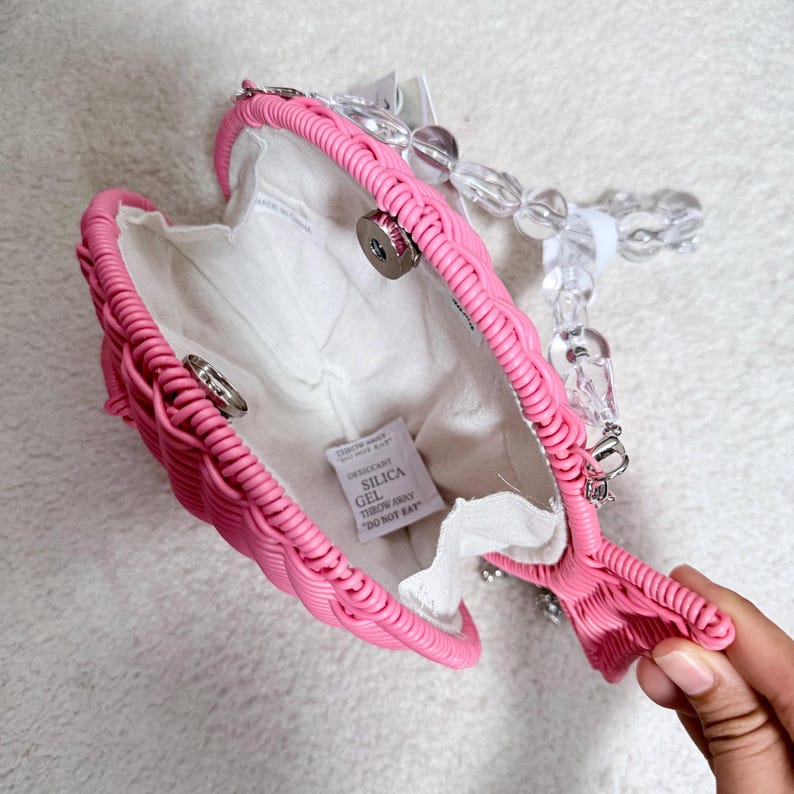 May include: A pink heart-shaped handbag with a woven design. The interior is white, and the handle is made of clear, rounded beads. The bag is open, revealing the inside. A small tag inside reads "SILICA GEL DO NOT EAT."