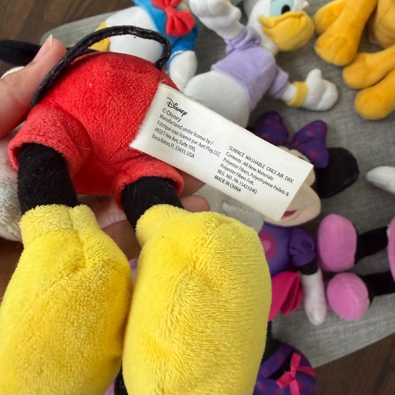 May include: A plush toy featuring red shorts and yellow shoes with a white fabric tag attached. The tag displays Disney branding, manufacturing information, and care instructions including Surface Washable Only, Air Dry, and Made in China text.
