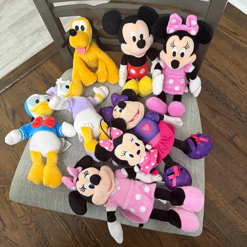 May include: A collection of Disney plush toys including Mickey Mouse, Minnie Mouse in various outfits, Donald Duck, Daisy Duck, and Pluto. The plush figures feature soft fabric, embroidered facial details, and iconic character clothing in bright colours.