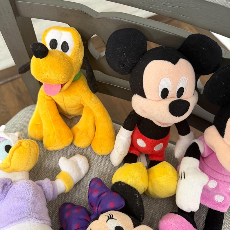 May include: A collection of plush toys featuring a yellow Pluto dog with a green collar, a black and white Mickey Mouse with red shorts and white buttons, a purple and white duck, and a Minnie Mouse with a purple polka dot bow and pink dress.