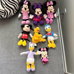 May include: A collection of eight plush toys featuring various cartoon characters. The set includes three Minnie Mouse dolls in pink and purple outfits, one Mickey Mouse, one yellow Pluto, one Donald Duck, one Daisy Duck, and one small Minnie Mouse.