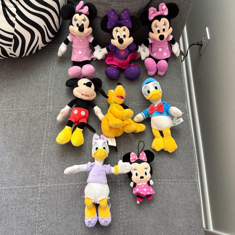 May include: A collection of eight plush toys featuring various cartoon characters. The set includes three Minnie Mouse dolls in pink and purple outfits, one Mickey Mouse, one yellow Pluto, one Donald Duck, one Daisy Duck, and one small Minnie Mouse.
